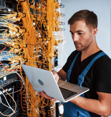 Young man in uniform and with laptop works with internet equipment and wires in server room.